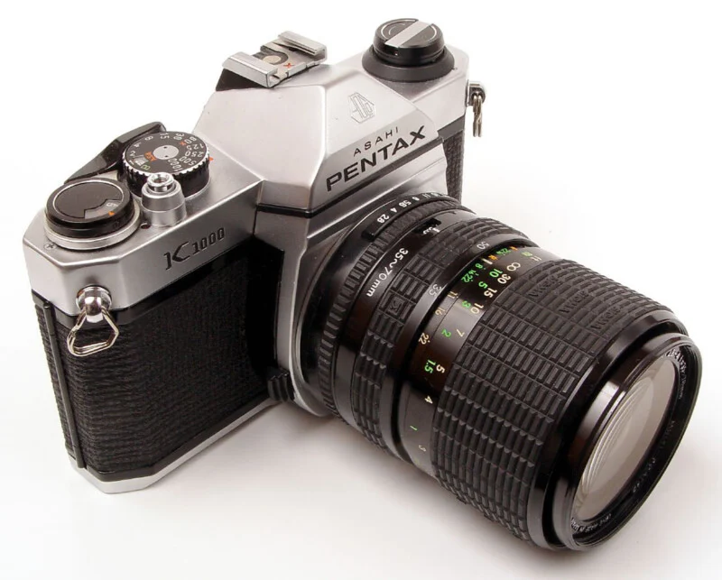 A silver Asahi Pentax K1000 35mm film SLR camera with a black 35-70mm zoom lens attached, viewed from a high angle against a white background.