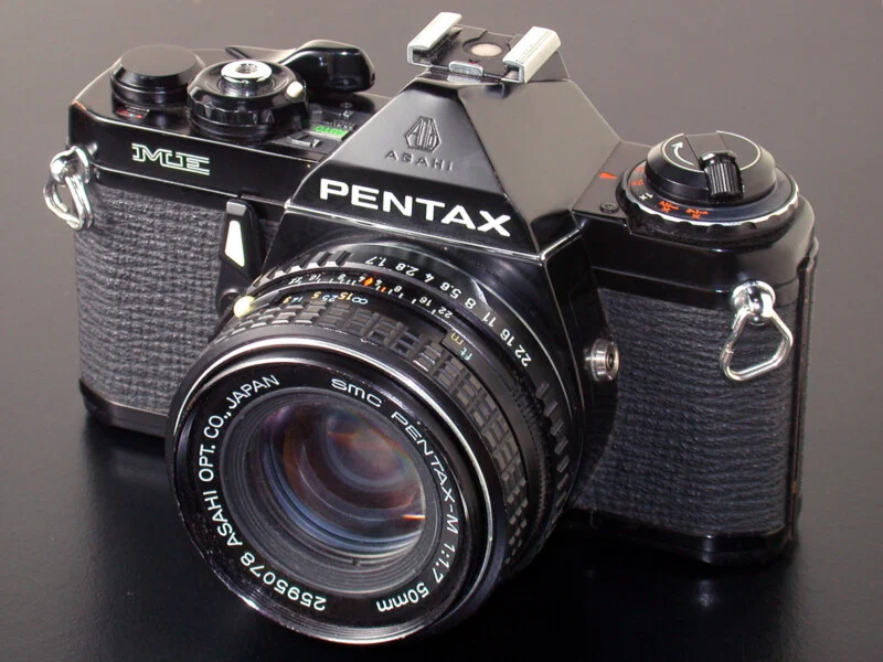 A black Pentax ME 35mm film camera with an SMC Pentax-M 50mm f/1.7 lens, positioned on a dark, reflective surface.