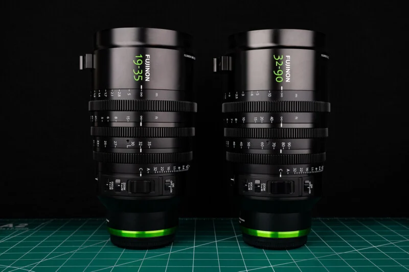 Two Fujinon cine zoom lenses, a 19-35mm and a 32-90mm, stand upright on a green cutting mat against a black background.