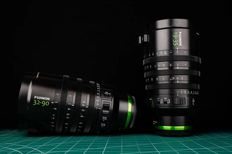 Two black Fujinon cinema zoom lenses, one lying horizontally and one standing vertically, sit on a green cutting mat against a black background.