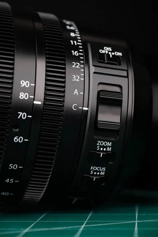 A close-up of a camera lens barrel featuring aperture settings, an OIS switch, a zoom toggle, and focus mode switches.