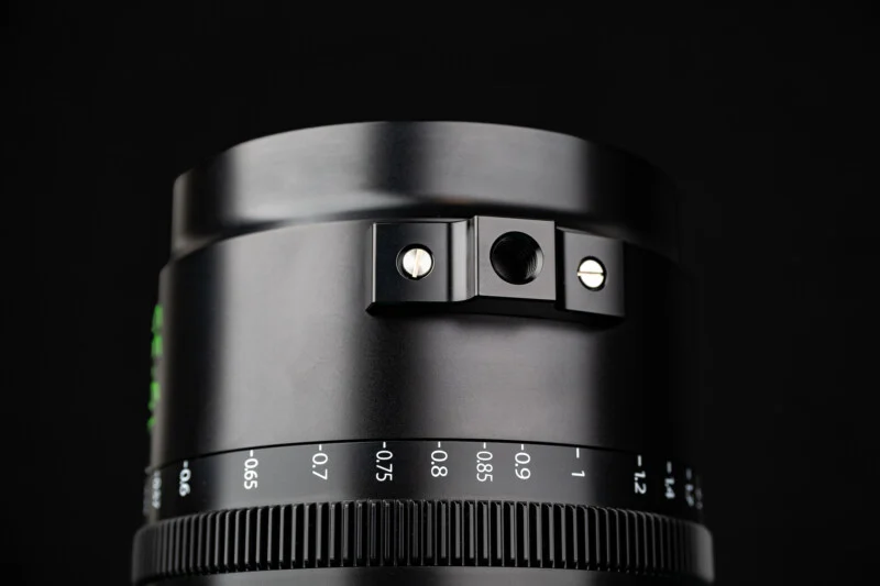 A close-up of a black camera lens barrel featuring a focus ring with distance markings and a mounting point with a threaded hole secured by two screws.