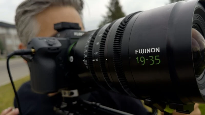 A person holds a professional camera with a large Fujinon 19-35 lens attached, focused on the lens barrel.