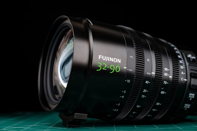 A black Fujinon 32-90 cinema zoom lens sits on a green cutting mat against a black background.