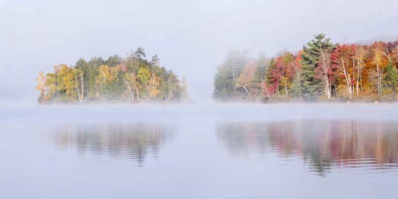 A calm lake reflects a misty shoreline featuring trees with autumn foliage in shades of yellow, orange, and red.