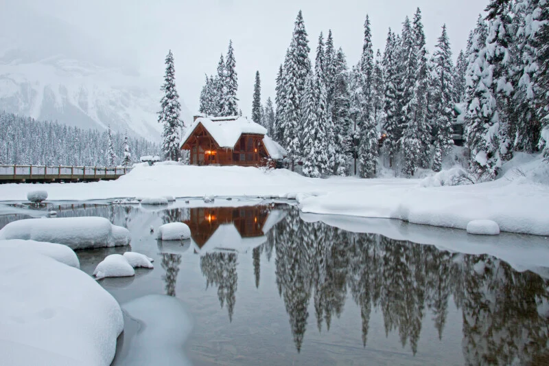 A wooden cabin sits on a snow-covered bank beside a calm, reflective pond surrounded by a dense forest of snow-laden pine trees.