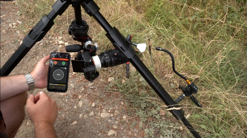 A person holds a smartphone displaying a lighting control app while standing next to a camera mounted on a tripod in a grassy field.