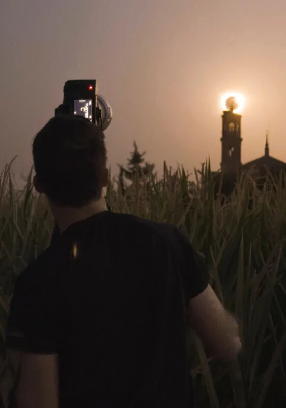 Een persoon in een zwarte shirt staat in een veld hoog gras en houdt een camera omhoog om de zonsondergang achter een kerktoren te fotograferen.