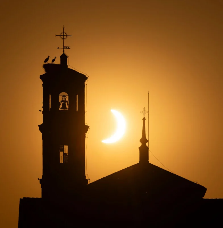 Een gedeeltelijke zonsverduistering verschijnt achter de silhouette van een kerkklokkentoren met een ooievaarsnest op de top en een kleinere aangrenzende toren.