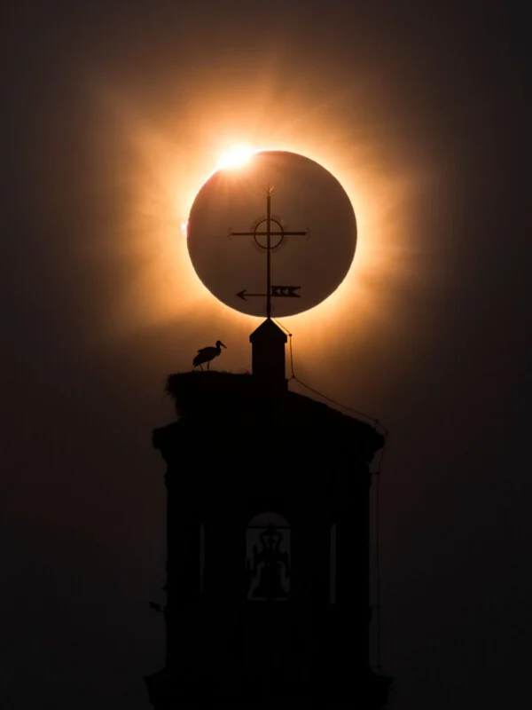 Zonsverduistering verschijnt direct achter de met een kruis bekroonde spits van een gebouw, waar een ooievaar in een nest staat.