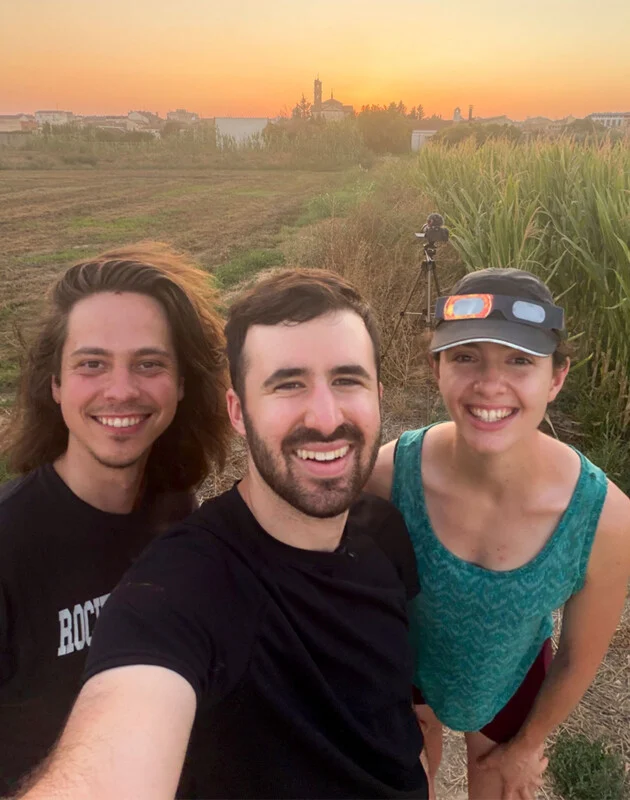 Drie mensen glimlachen voor een selfie in een veld bij zonsondergang, met een camera op een statief op de achtergrond.