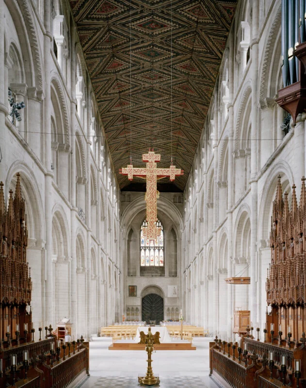 The interior of a long, stone cathedral nave features a high, patterned wooden ceiling, a large suspended cross, and rows of wooden pews leading to an altar.