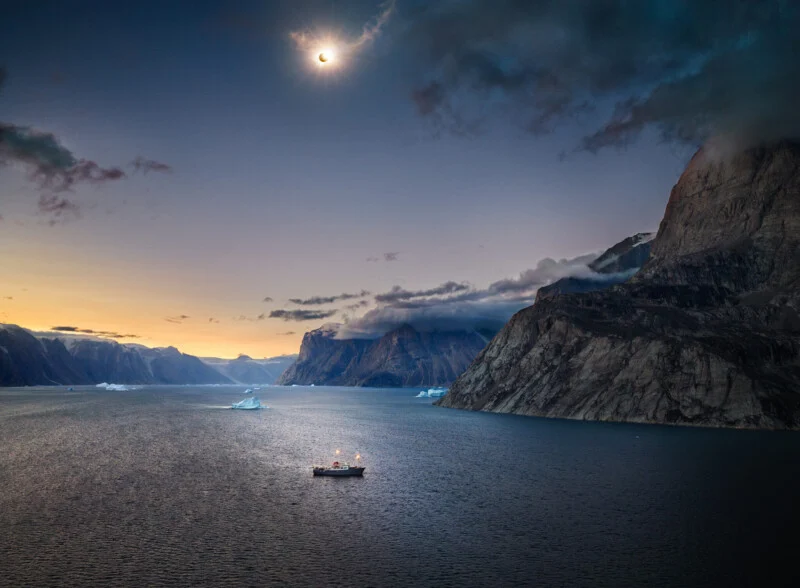 A small boat floats on a calm, dark body of water surrounded by steep, rocky mountains under a solar eclipse in the sky.
