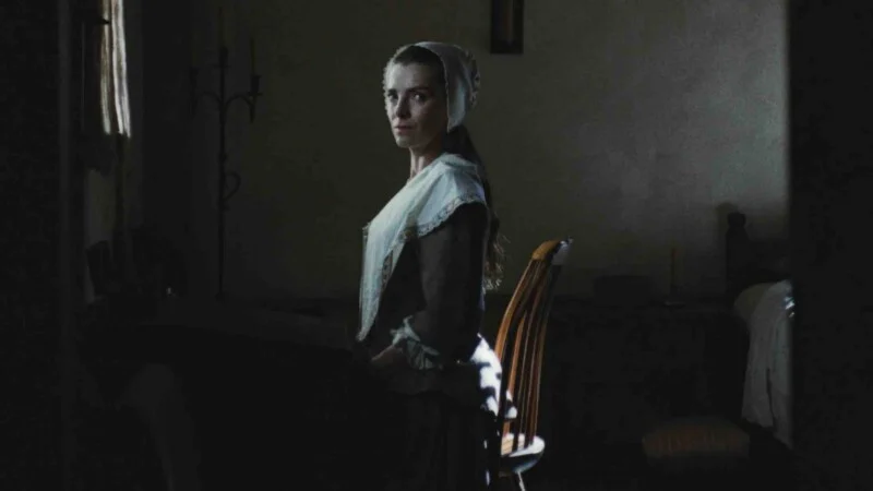 A woman in a historical white cap and shawl stands in a dimly lit room, looking back over her shoulder toward the camera.
