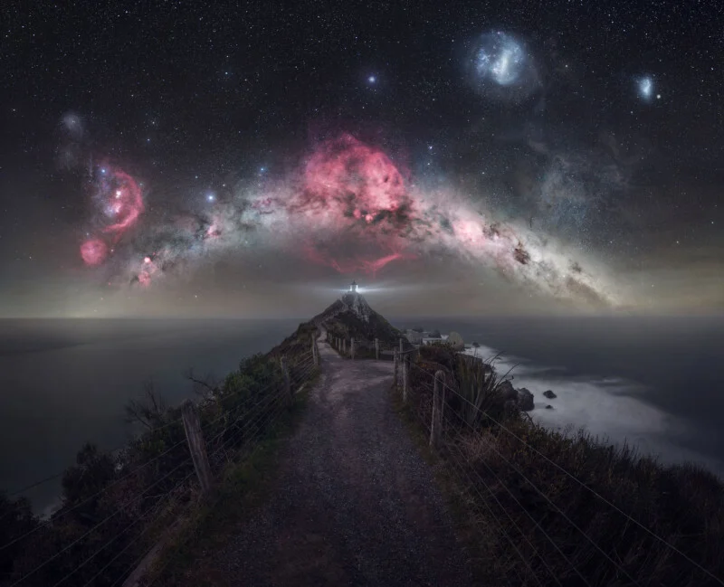 A lighthouse sits at the end of a narrow, fenced path overlooking the ocean beneath a vibrant, star-filled night sky.
