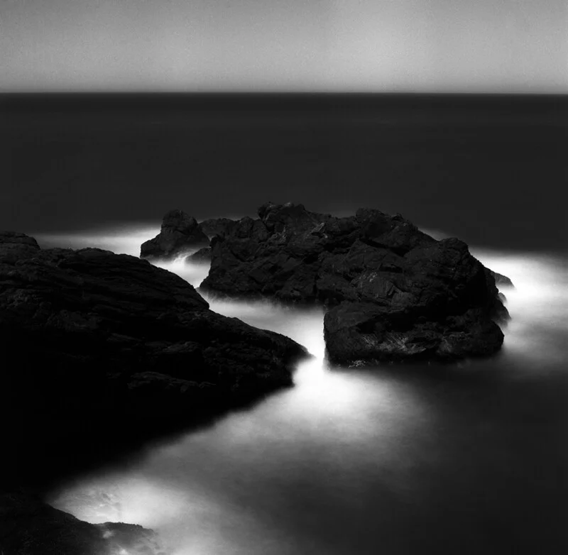 A long-exposure, black-and-white photograph shows dark, jagged rocks surrounded by blurred, glowing white sea foam.