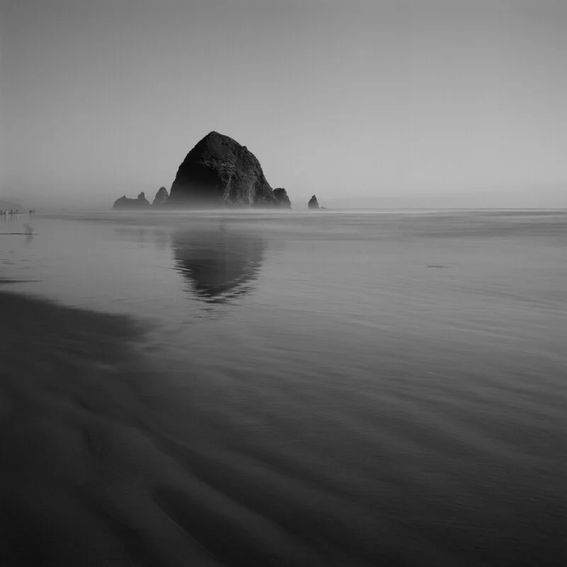A large, dark sea stack stands in the distance on a flat, sandy beach, reflected in the calm, shallow water at low tide.