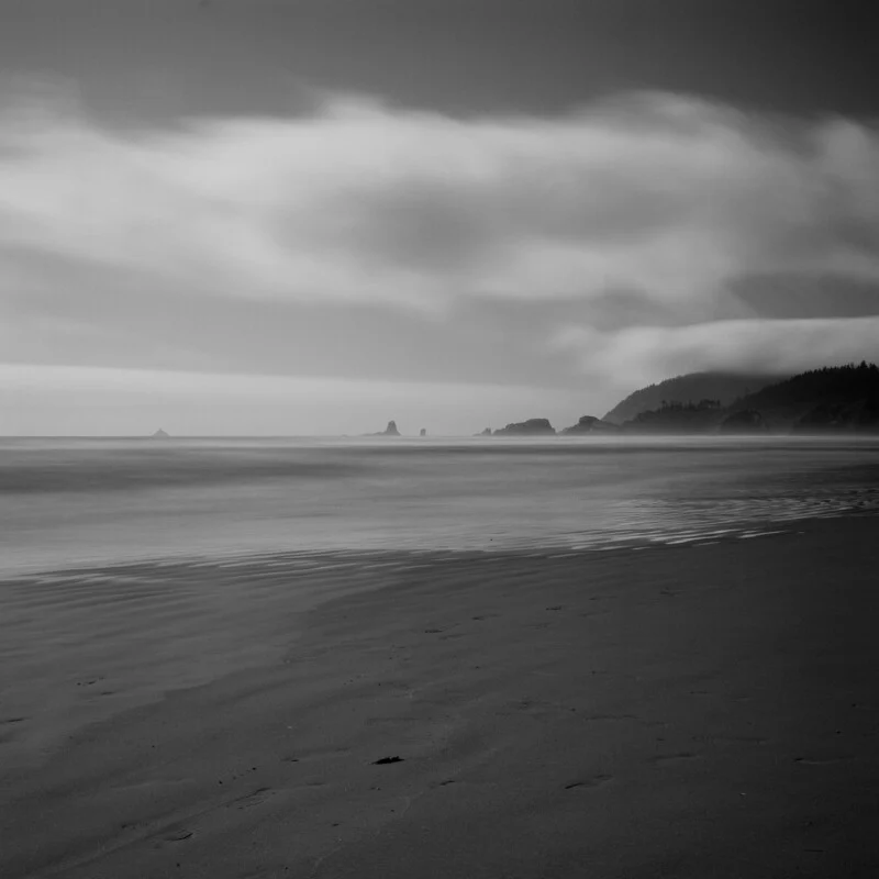 A black and white long-exposure view of a sandy beach with calm water, distant rock formations, and low-hanging clouds.