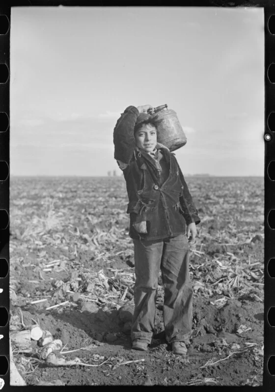 A young boy in a worn, dark corduroy jacket stands in a harvested field, carrying a metal water jug on his shoulder.