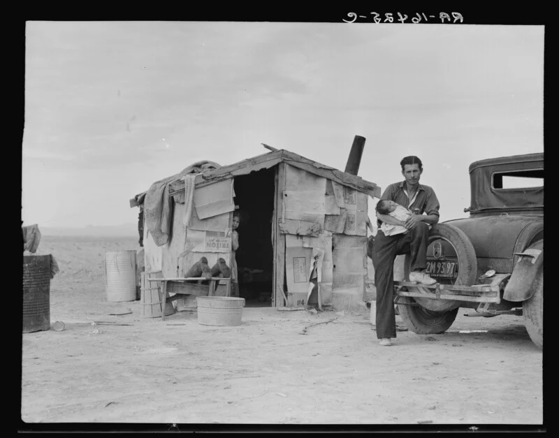 A man holds an infant while leaning against a car parked next to a small, makeshift shack constructed from cardboard and wood, with a child peering from the doorway.