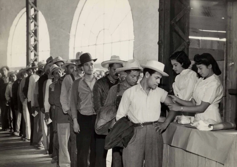 A line of men wearing hats and work clothes wait to receive vaccinations from two nurses at a table.