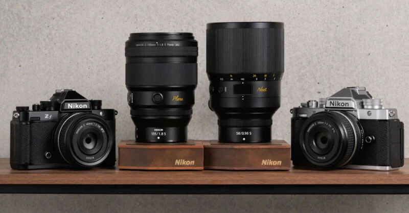 Two Nikon Zf cameras, one black and one silver, are positioned on a wooden shelf alongside two large Nikon lenses, the 135mm Plena and 58mm Noct, each resting on a branded wooden stand.