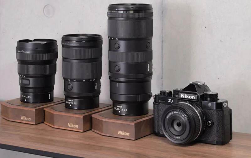 Three Nikon lenses on wooden stands sit next to a Nikon Zf camera with a 40mm lens on a wooden surface.