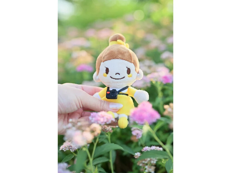 A hand holds a small plush doll with brown hair in a bun, wearing a yellow dress and a tiny black camera, amidst pink flowers.