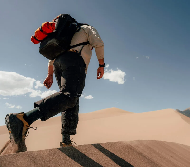 A hiker with a large backpack walks across the crest of a sand dune under a clear blue sky.