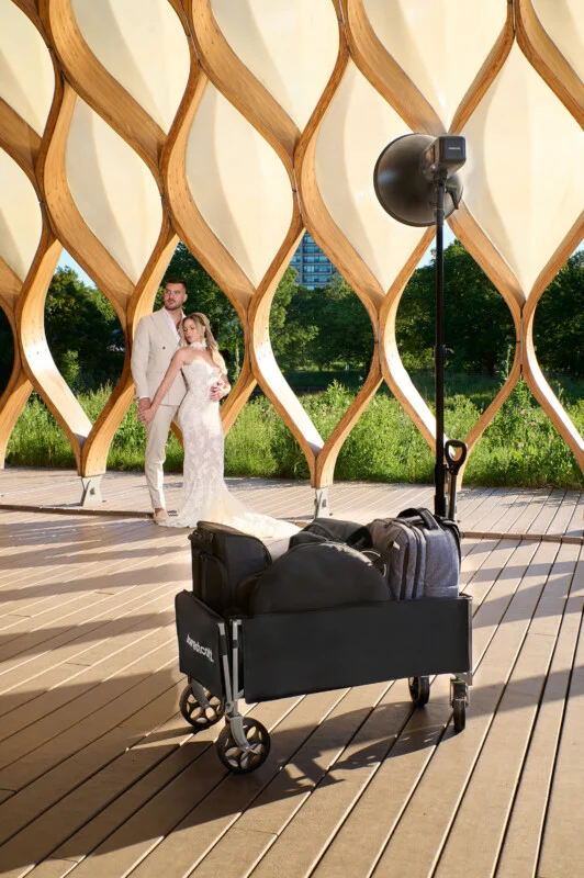 A bride and groom stand together under a wooden pavilion while a photography equipment cart with a light stand sits in the foreground.