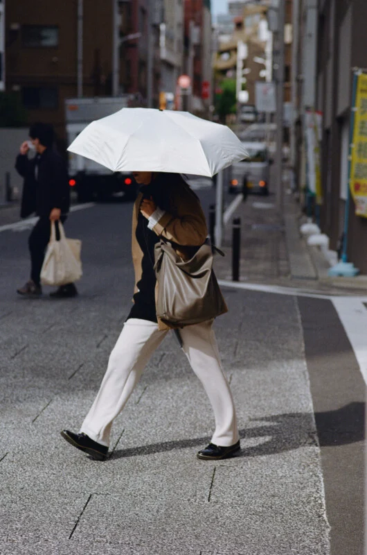 A person walks across a crosswalk while holding a white umbrella, wearing a tan coat, black top, and white trousers with a large grey shoulder bag.