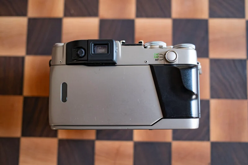 A silver and black film camera sits on a wooden chessboard, viewed from the back, showing the viewfinder, a focus mode selector, and a black grip.