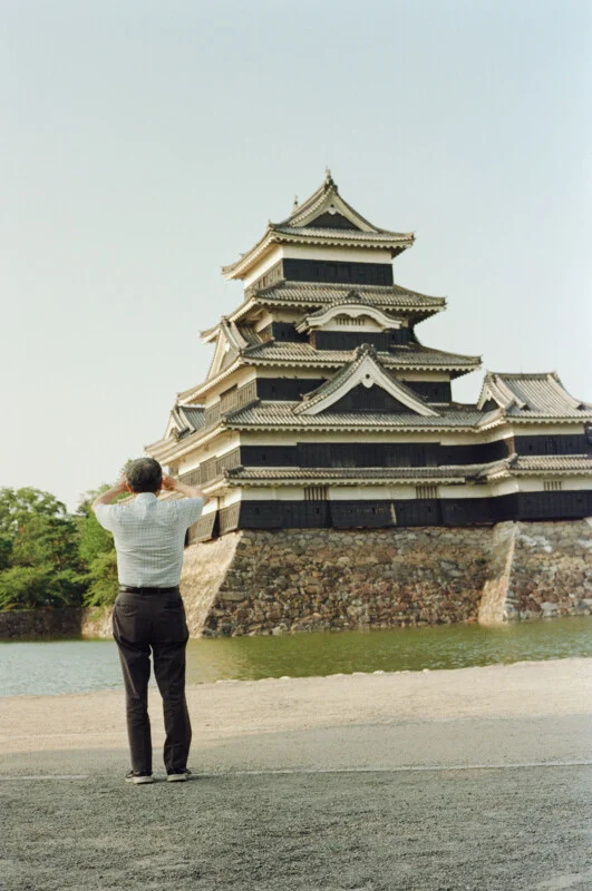 A man in a light-colored short-sleeved shirt and dark trousers stands with his hands raised to his head, looking at a large, multi-storied black castle across a moat.
