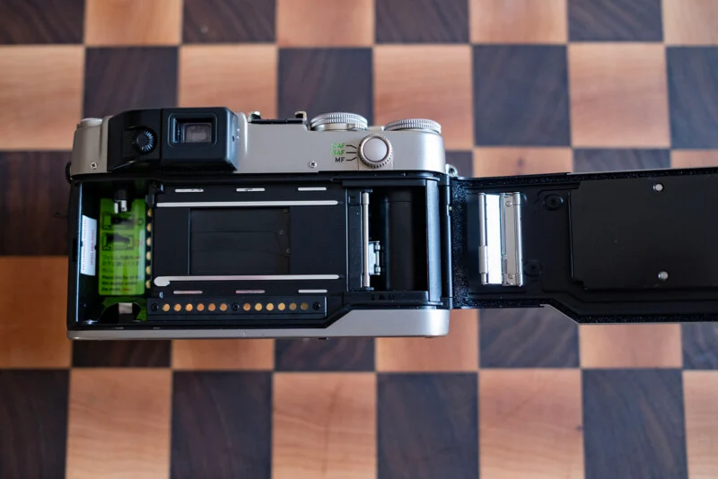 An open 35mm film camera sits on a wooden checkerboard surface, showing the film chamber, shutter curtains, and take-up spool.