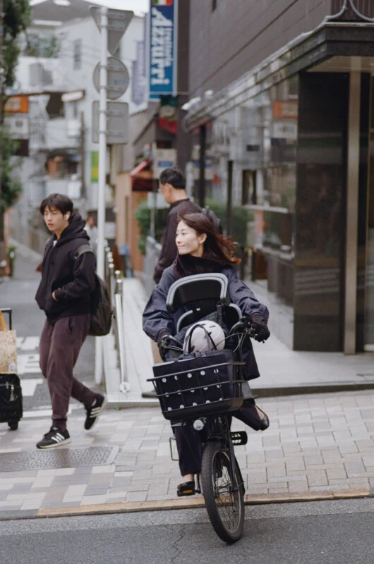 A woman rides a bicycle with a child seat and a basket through a city street, while a young man walks past her in the background.