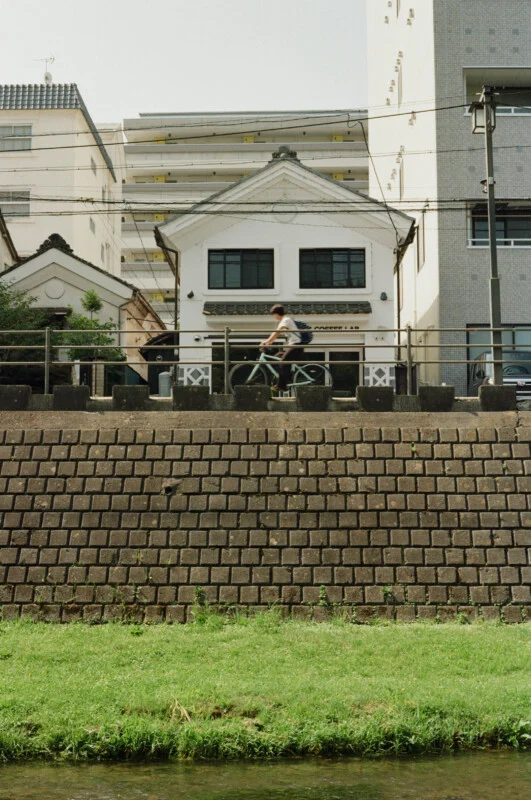 A person rides a bicycle along a path in front of a white building with a gabled roof, situated above a stone retaining wall.