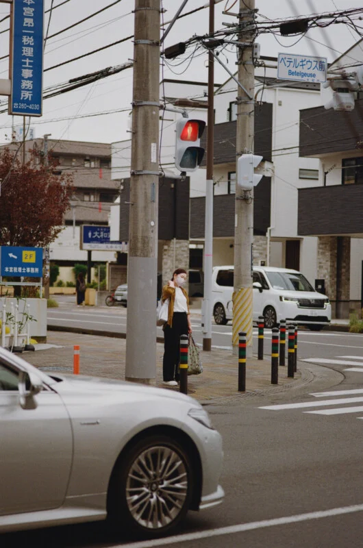 A person wearing a face mask stands on a sidewalk corner next to a traffic light pole, with a silver car in the foreground.