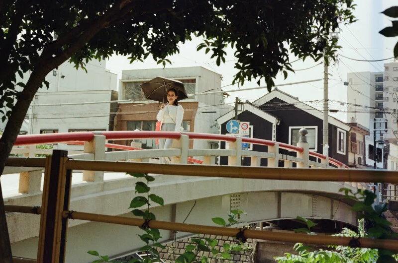 A woman holding a black umbrella stands on a white bridge with red railings, with buildings and utility poles in the background.