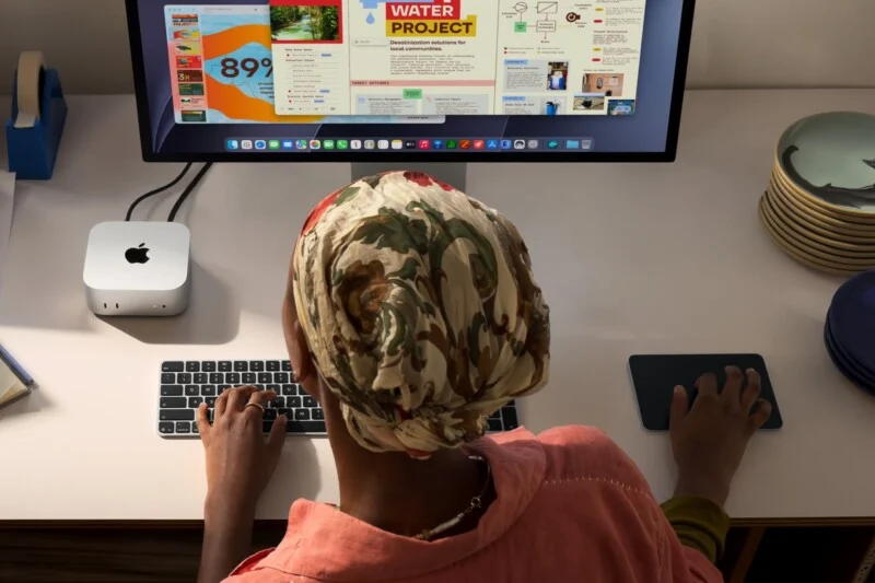 Een persoon met een bloemige hoofdoek zit aan een bureau en gebruikt een Mac Mini, een toetsenbord en een trackpad om een waterprojectpresentatie op een groot monitor te bekijken.