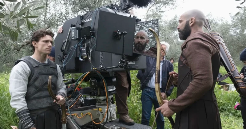 Actors in medieval costumes stand near a large film camera outdoors on a grassy set. Crew members adjust equipment while one actor holds a bow and another has arrows on his back.