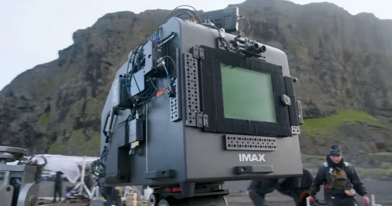 A large IMAX camera is set up outdoors with rocky hills in the background. Film crew members and equipment are visible around the scene.