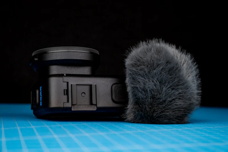 Een zwarte actiecamera met een ronde lensfilter naast een pluizige grijze windgeluidmuur op een blauwe rasterplaat tegen een zwarte achtergrond.