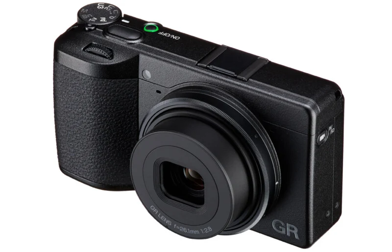 A black Ricoh GR IIIx digital camera angled to show the front and top, featuring a fixed 26.1mm f/2.8 lens and a mode dial.