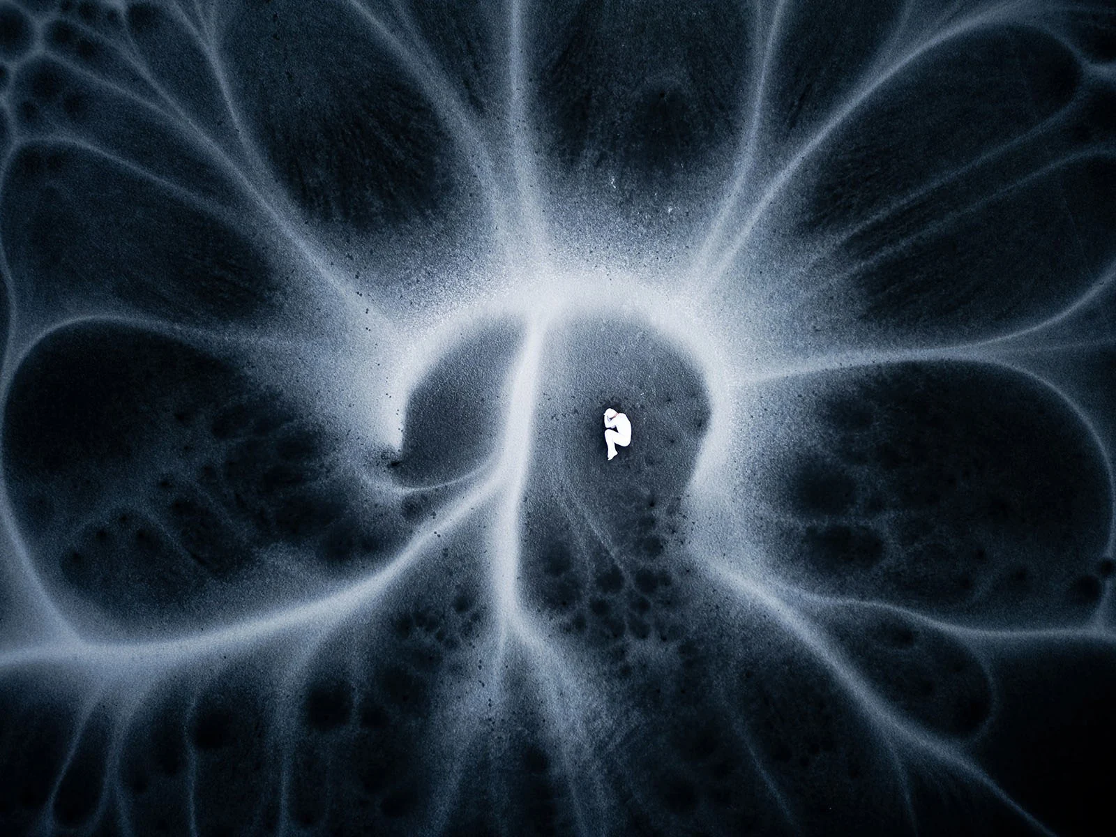 A small, white, fetal-shaped figure lies in the center of a dark, swirling, abstract pattern resembling a nebula or fluid.