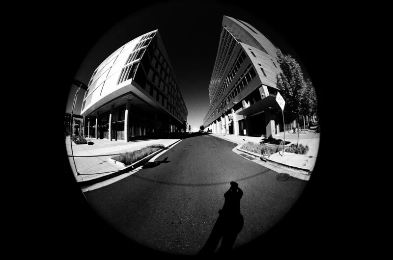 A fisheye, black-and-white view of a street between two modern buildings, with the photographer's shadow cast on the asphalt.