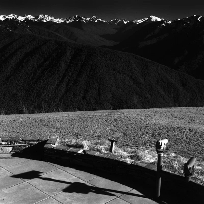 A black-and-white view from a stone-walled overlook featuring two coin-operated binoculars, a small sign, and a vast mountain range in the distance.