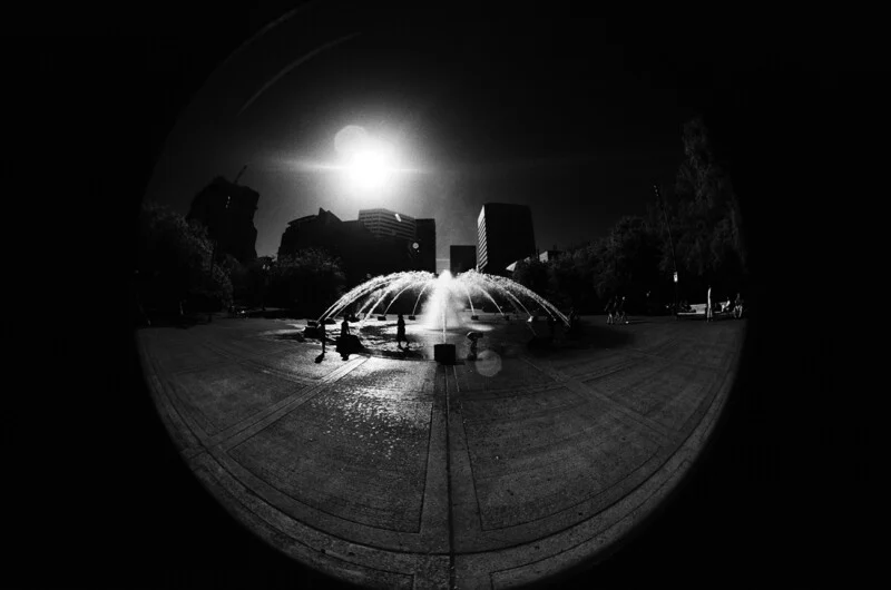A black and white fisheye view of a circular fountain spraying water in a city plaza under a bright sun.