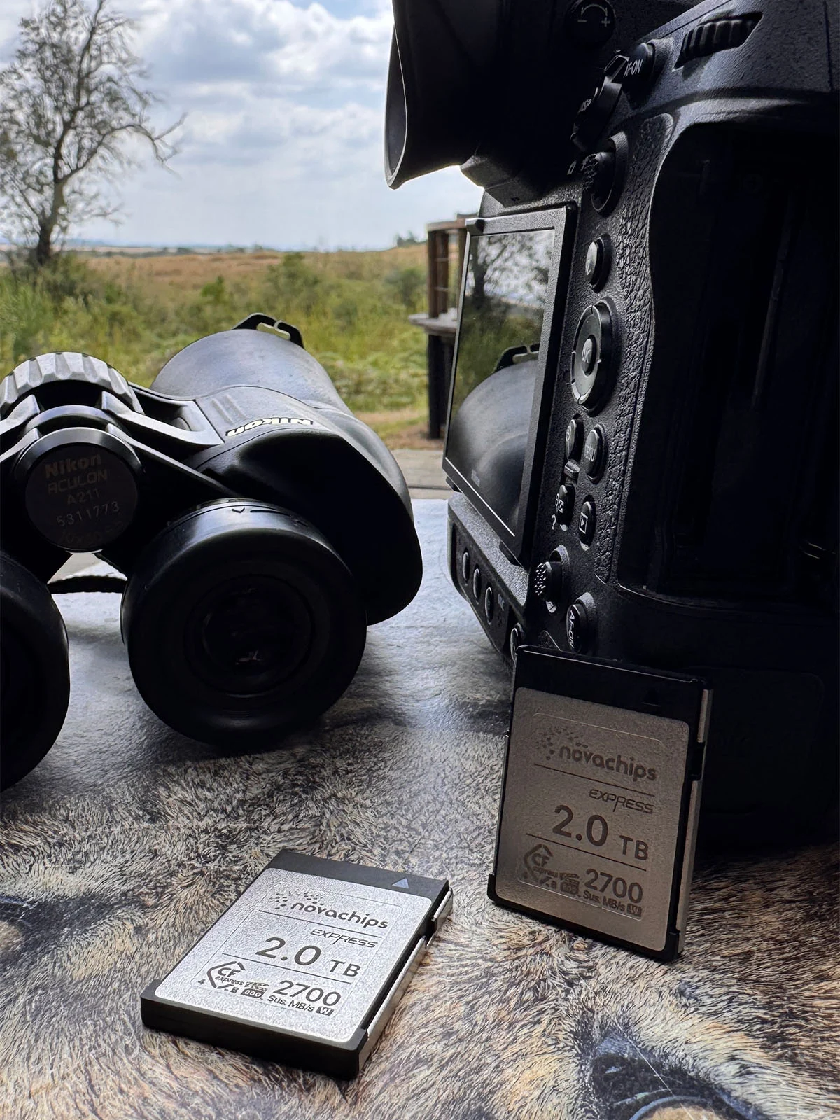 Two Novachips 2.0 TB CFexpress memory cards sit on a textured surface next to a Nikon camera and Nikon Aculon binoculars.