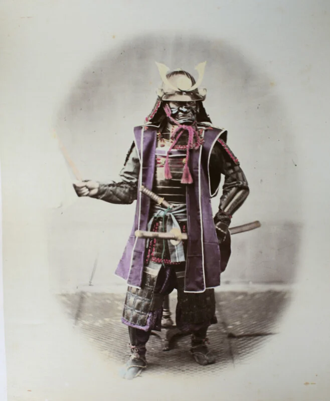 A hand-colored studio portrait of a samurai in full armor, wearing a horned helmet and a fierce mask, holding a baton.