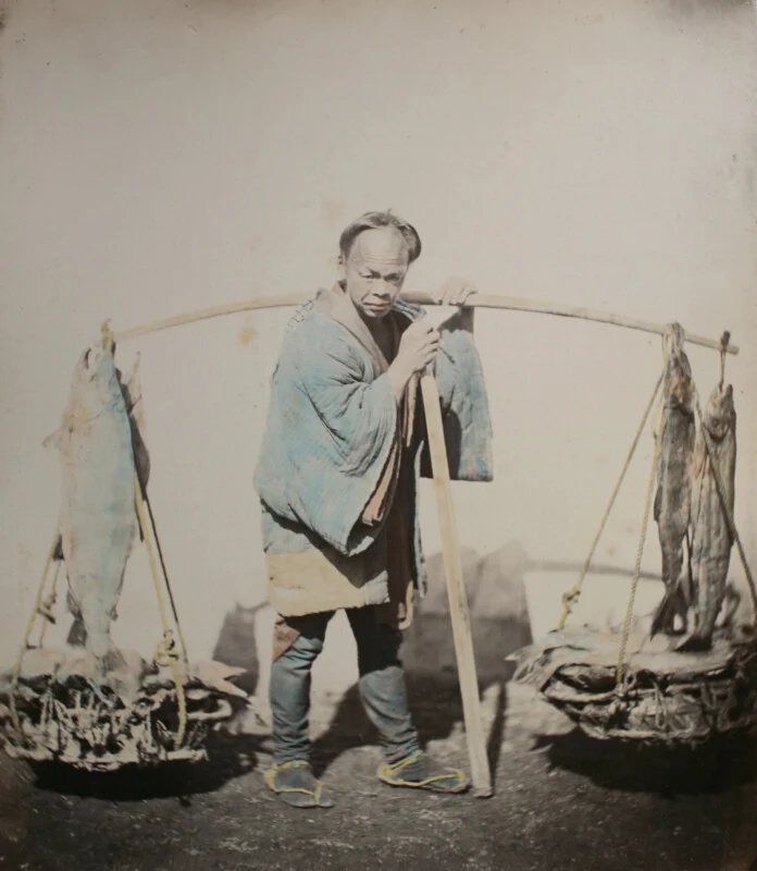A man in a blue kimono carries a bamboo pole across his shoulders with baskets of fish hanging from each end.
